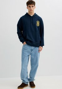 Joven de pie con una sudadera con capucha azul marino con logotipo amarillo, vaqueros azul claro y zapatillas marrones sobre un fondo claro y liso.