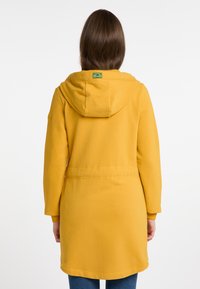 Felpa gialla con cappuccio e cordino, maniche lunghe e una cintura in vita. Materiale morbido con etichetta sul retro. Texture liscia.