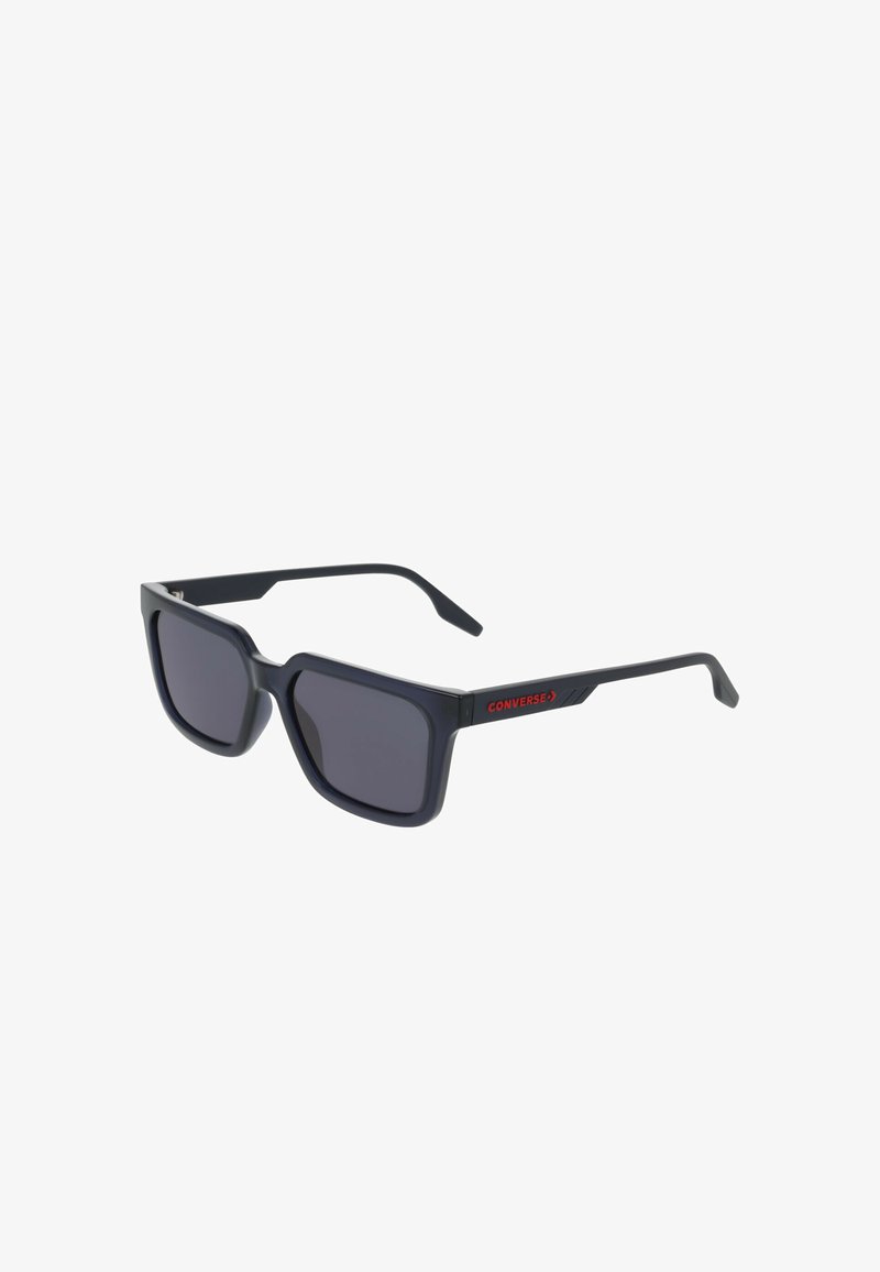 Lunettes de soleil en plastique noir avec de grandes lentilles carrées, verre teinté foncé, et un logo rouge « CONVERSE » sur le côté de la monture.