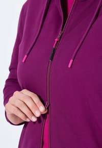 Sweat à capuche zippé violet en tissu doux avec des détails de fermeture éclair et de cordons contrastants roses, présenté dans un design ajusté et une texture lisse.