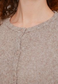 Vrouw die een zachte, beige gebreide cardigan met knopen en een ronde hals draagt, waarbij een deel van haar nek en krullend haar zichtbaar is.