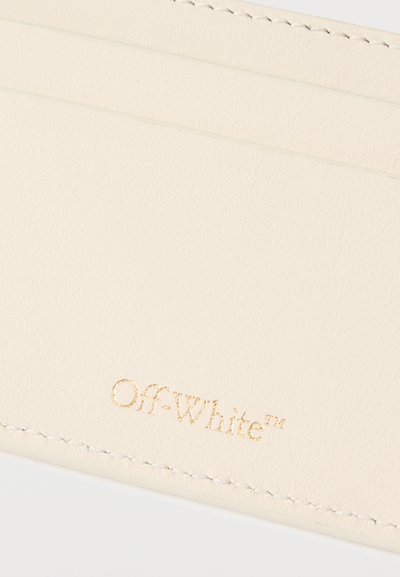 Εγγύς λήψη ενός μπεζ δερμάτινου θηκίου καρτών με ανάγλυφο χρυσό λογότυπο "Off-White™" κοντά στις ραμμένες άκρες.