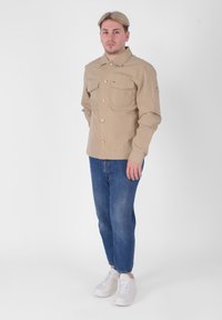 Camicia beige con bottoni, dotata di due tasche sul petto, realizzata in tessuto leggero. Abbinata a jeans in denim blu e sneakers bianche.