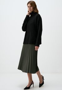 Schwarzer Strickpullover mit Rollkragen kombiniert mit einem plissierten olivgrünen Midirock und schwarzen High-Heels. Schlichte, strukturierte Kleidungsdetails.