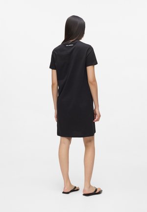 Kobieta z długimi ciemnymi włosami ubrana w czarną sukienkę o długości do kolan z krótkimi rękawami i czarne sandały, stojąca tyłem na jednolitym białym tle.