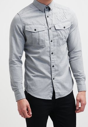 Homme portant une chemise cintrée gris clair à boutons avec deux poches poitrine et un pantalon noir sur un fond uni.