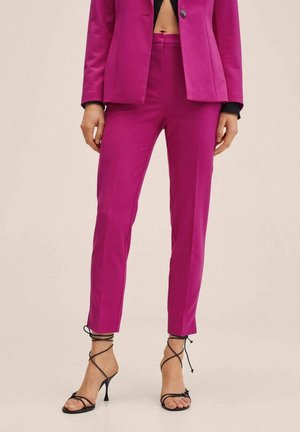 Vrouw draagt felroze op maat gemaakte broek en bijpassende blazer, gecombineerd met zwarte sandalen met hak en bandjes tegen een effen achtergrond.