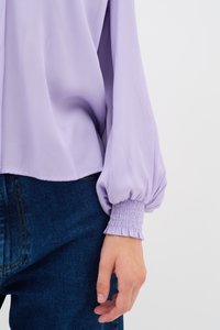 Blusa lilla con maniche a sbuffo, caratterizzata da un polsino arricciato. Indossata con jeans in denim blu, evidenziando un tessuto morbido e leggero.