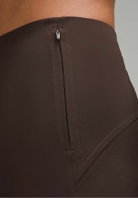 Gros plan sur une personne portant des leggings marron taille haute avec un détail de fermeture éclair latérale verticale près de la hanche.