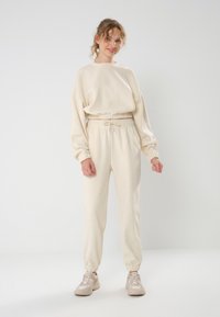 Cremefarbener Trainingsanzug mit lockerer Passform, der eine weiche Textur, einen elastischenBund und Bündchen aufweist. Kombiniert mit hellbeigen Sneakers.