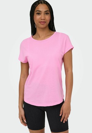 Woman standing, wearing a light pink short-sleeve t-shirt and black fitted shorts, against a plain light background.