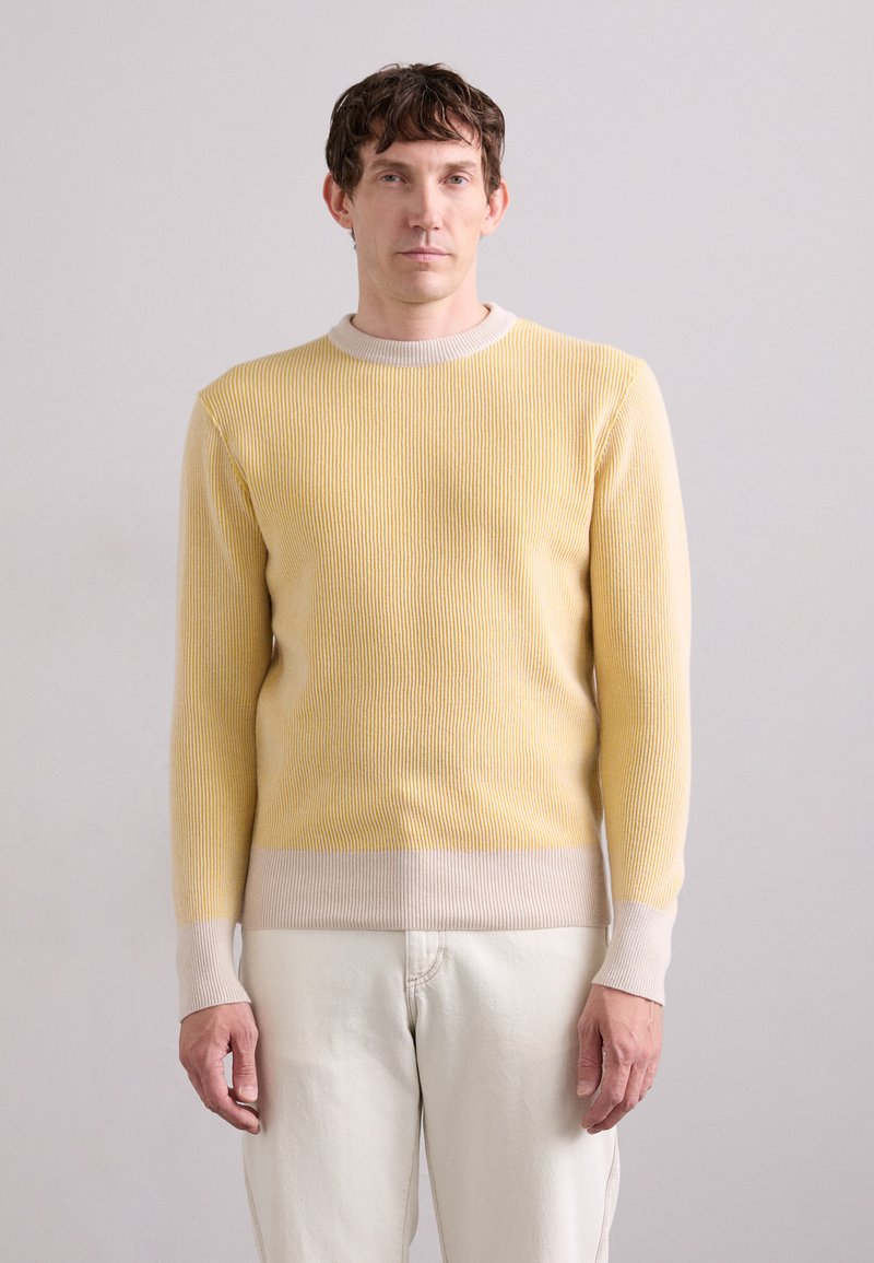 Gelber gerippter Pullover mit einem hellbeigen Saum und Ärmeln. Verfügt über lange Ärmel und einen klassischen Rundhalsausschnitt. Getragen mit hellen Hosen.