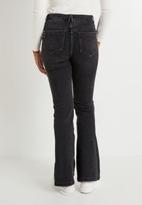 Persona con pantalones vaqueros negros ajustados con bolsillos traseros y zapatillas blancas, de pie contra un fondo blanco liso.