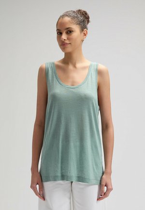 Donna con capelli ricci raccolti in uno chignon, indossa orecchini a cerchio dorati, un top verde senza maniche e pantaloni bianchi, con uno sfondo grigio semplice.