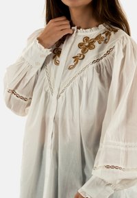 Blusa bianca in cotone con un collo alto arricciato, modelli floreali ricamati in marrone, chiusure a bottone e delicati strati di pizzo sulle maniche.
