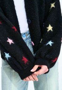 Ruky zložené vpredu, oblečené v čiernom huňatom kardigáne s farebnými vzormi hviezd a modré džínsy s viditeľnými gombíkmi.