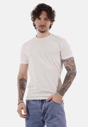 Uomo con capelli ricci, indossa una t-shirt bianca semplice e jeans blu, mostra tatuaggi dettagliati su entrambe le braccia, in piedi con una mano in tasca.