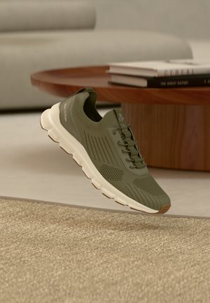 Sneaker verde oliva lavorata a maglia con suola bianca e linguetta, posizionata vicino a un tavolino di legno con libri impilati su un tappeto beige.
