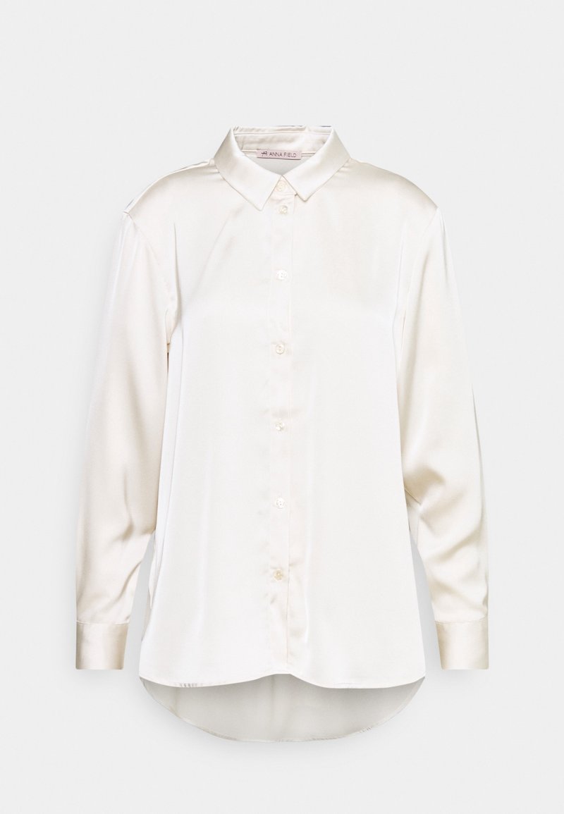 Blouse blanche en satin à manches longues avec col pointu, boutons sur le devant et ourlet légèrement arrondi, présentée sur un fond clair uni.