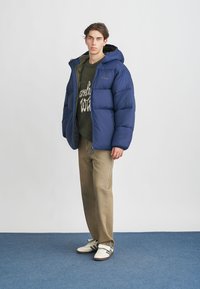 Blue puffer jacket with a hood, featuring a quilted design. Worn over a green sweater and beige pants; paired with white sneakers.