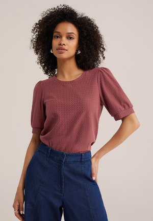 Femme aux cheveux bouclés portant un haut texturé mauve à manches courtes et un jean taille haute à jambes larges bleu foncé, posant la main sur la hanche.