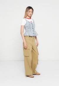 Denim peplumtop met knoopdetail, gelaagd over een witte T-shirt, gecombineerd met brede beige cargobroek en lichtbruine schoenen.