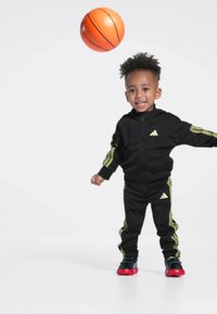 Black athletic tracksuit with yellow stripes, featuring an adidas logo. Child in sneakers dribbling an orange basketball, smiling.