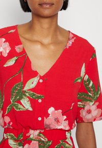 Vestido floral vermelho com decote em V, mangas curtas e frente com botões. O tecido exibe grandes flores cor-de-rosa e folhas verdes.