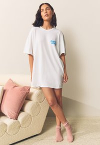 Vestido camiseta blanco con el gráfico "The Sunday Brunch" en azul. La modelo lleva calcetines rosa acanalados. Corte suave y relajado con textura de algodón lisa.