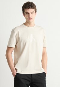 Beige cotton t-shirt featuring a white CK logo, round neckline, short sleeves, and a relaxed fit, paired with black pants.