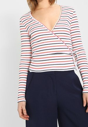 Kobieta ubrana w białą, długorękawową bluzkę typu wrap z czerwonymi, czarnymi i niebieskimi poziomymi paskami, w połączeniu z ciemnoniebieskimi, wysokim stanem spodniami.