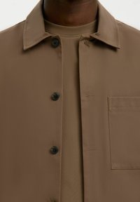 Gros plan sur une personne portant une veste marron à boutons par-dessus un t-shirt assorti marron à col rond, montrant le col, les boutons et la poche poitrine.