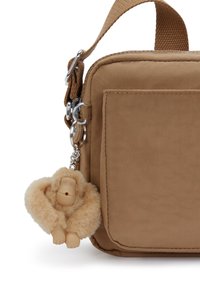 Beige taske med tværrem, der har en sølvfarvet kæde-nøglering med en blød abe-charme. Inkluderer en frontlomme og en solid rem.