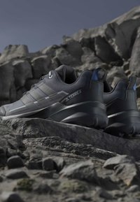 Ζεύγος σκούρων γκρι παπουτσιών trail running Adidas Terrex σε πετρώδες έδαφος με τραχιές ορεινές πέτρες στο φόντο κάτω από καθαρό ουρανό.