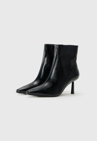 Une paire de bottines noires en cuir lisse à bout pointu avec des talons aiguilles fins de hauteur intermédiaire sur un fond clair.
