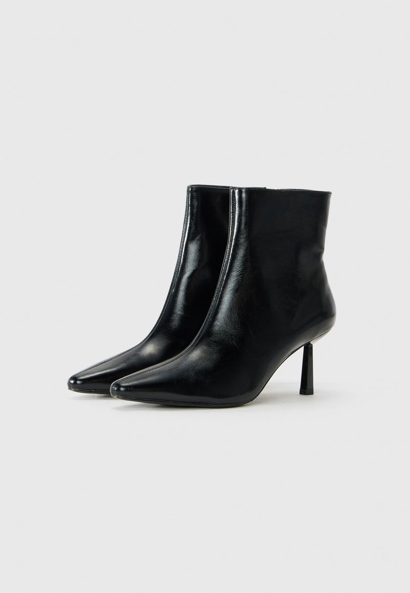 Un paio di eleganti stivali alla caviglia in pelle nera lucida con punta affusolata e tacchi a stiletto slim di medio altezza su uno sfondo chiaro e neutro.