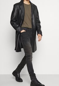 Persona caminando con un abrigo de cuero negro, camisa verde oliva, jeans negros con textura y botas negras de cordones sobre un fondo liso.