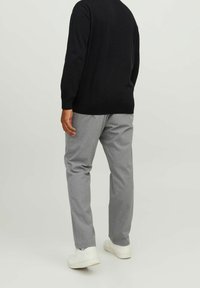 Personne portant un pantalon gris à fines rayures, des baskets blanches et un haut à manches longues noir, debout devant un fond clair uni.