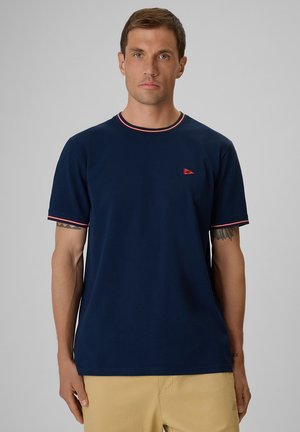 Mężczyzna z krótkimi brązowymi włosami w granatowym t-shircie z czerwonym obszyciem i beżowych spodniach, na jednolitym szarym tle.