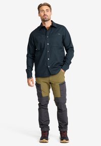 Man som står och bär en svart skjorta med knappar, tvåtoniga gröna och mörkgrå cargo-byxor samt svarta vandringsskor med orange snören.