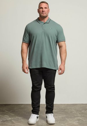Man met een gespierd postuur, gekleed in een groen poloshirt, zwarte broek en witte sneakers, staand tegen een effen achtergrond.
