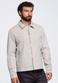 Homme avec barbe portant une veste zippée gris clair et un pantalon foncé, regardant légèrement vers la droite sur un fond clair uni.