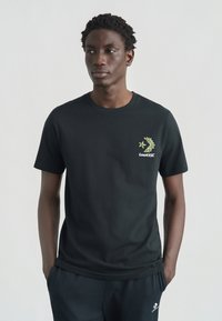 Μαύρο βαμβακερό μπλουζάκι με πράσινο λουλουδάτο σχέδιο και την επιγραφή "Converse" στο αριστερό στήθος. Κοντά μανίκια και στρογγυλή λαιμόκοψη.