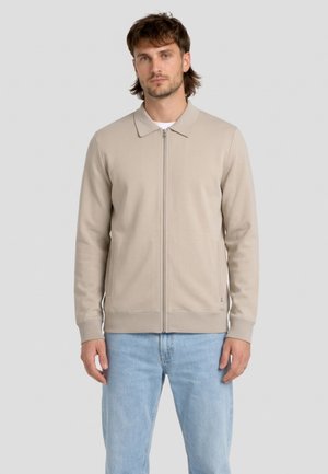 Man die een beige jas met rits en kraag draagt en een lichtblauwe spijkerbroek, staand tegen een effen lichtgrijze achtergrond.