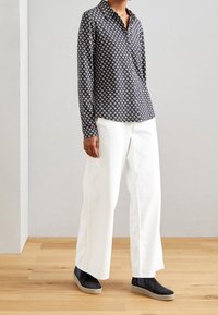Personne portant une chemise à manches longues noire à motifs, un large pantalon blanc et des chaussures noires à enfiler, se tenant sur un sol en bois contre un mur gris.