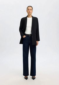 Kvinne står med svart blazer, hvit topp, mørkeblå jeans, svart belte og svarte spissesko mot en lys ensfarget bakgrunn.