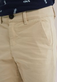 Beige bomulls chinos med en mjuk struktur, framfickor och knappstängning. Tyget visar subtila veck och har en klassisk passform.