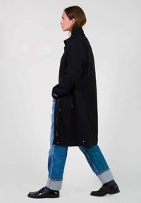Manteau long noir en laine, avec un col haut, des poches latérales et une silhouette en A, associé à un jean bleu et des chaussures noires.
