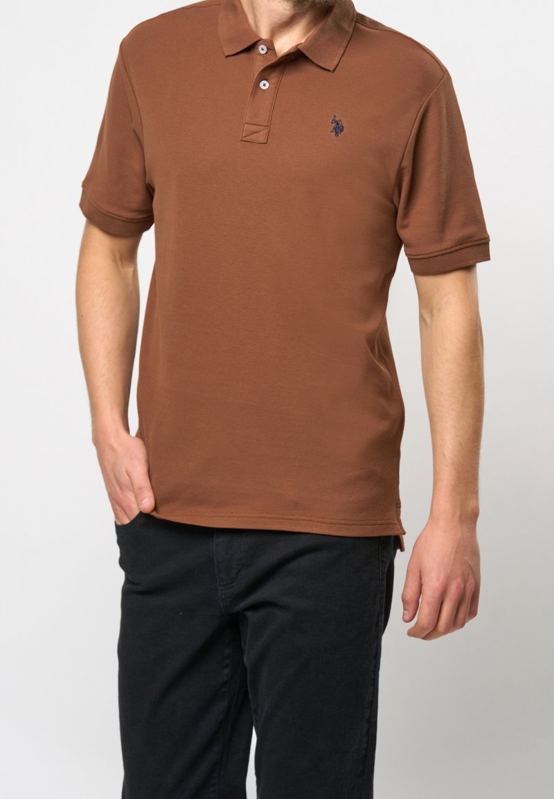 Homme portant un polo marron à manches courtes avec des boutons et un pantalon noir, debout avec une main dans la poche et l’autre détendue.