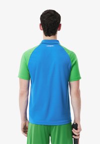 Blå och grön polo skjorta med korta ärmar och krage. Baksidan har Lacoste-logotypen nära nacken. Texturerad tyg med kontrasterande färger.